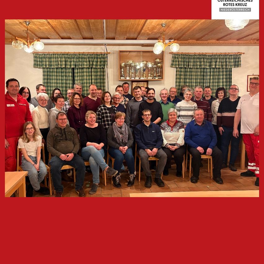 Eine Gruppe von Menschen sitzt auf Stühlen in einem Raum mit Holzdecke, ausgestellten Trophäen und rotem Hintergrund. Einige tragen Brillen, Schals und Turnschuhe.