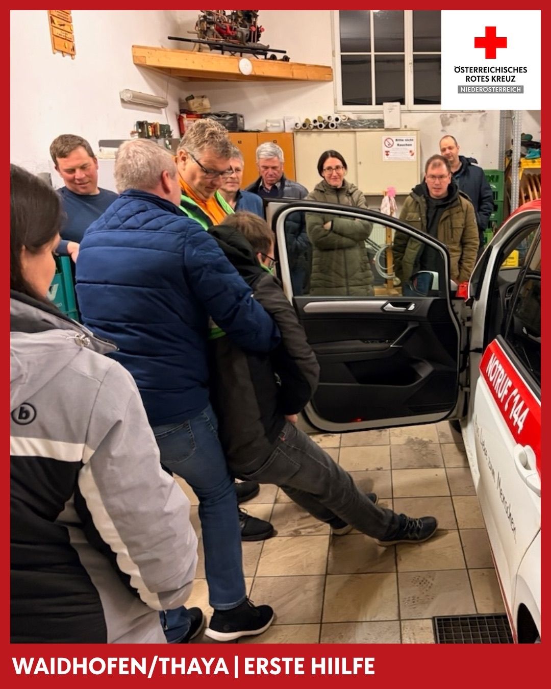 In einer Werkstatt versammeln sich mehrere Personen um ein Auto mit offener Tür. Ein Mann wird von einem anderen Mann mit Brille unterstützt. Eine Frau mit Brille beobachtet in der Nähe. Das Auto ist mit der Nummer 144 gekennzeichnet.