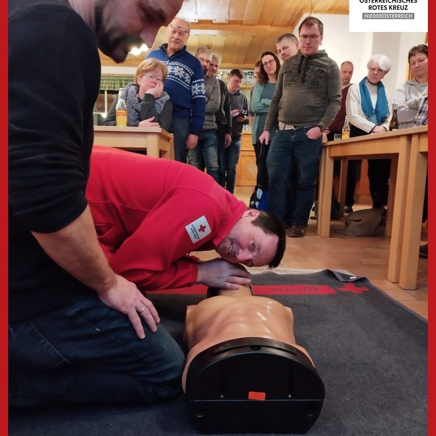 Ein Mann übt CPR an einem Mannequin, während Zuschauer zusehen, einige sitzen und andere stehen, in einem Raum mit Holztischen und einem roten Rand.