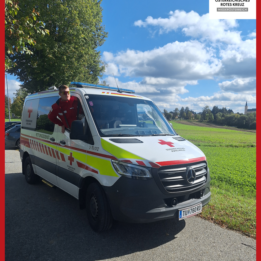 Ein Mann steht neben einem Krankenwagen mit rotem Kreuz-Symbol an einem sonnigen Tag. Bäume und eine Rasenfläche sind im Hintergrund zu sehen. Der Krankenwagen ist am Straßenrand geparkt.