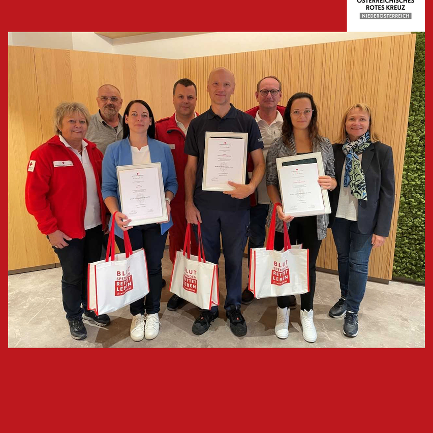 Seven people pose for a photo while holding certificates and bags. They are standing in front of a wooden wall. The bags have the text 'Blut spenden retten leben'. The man in the middle holds a certificate.