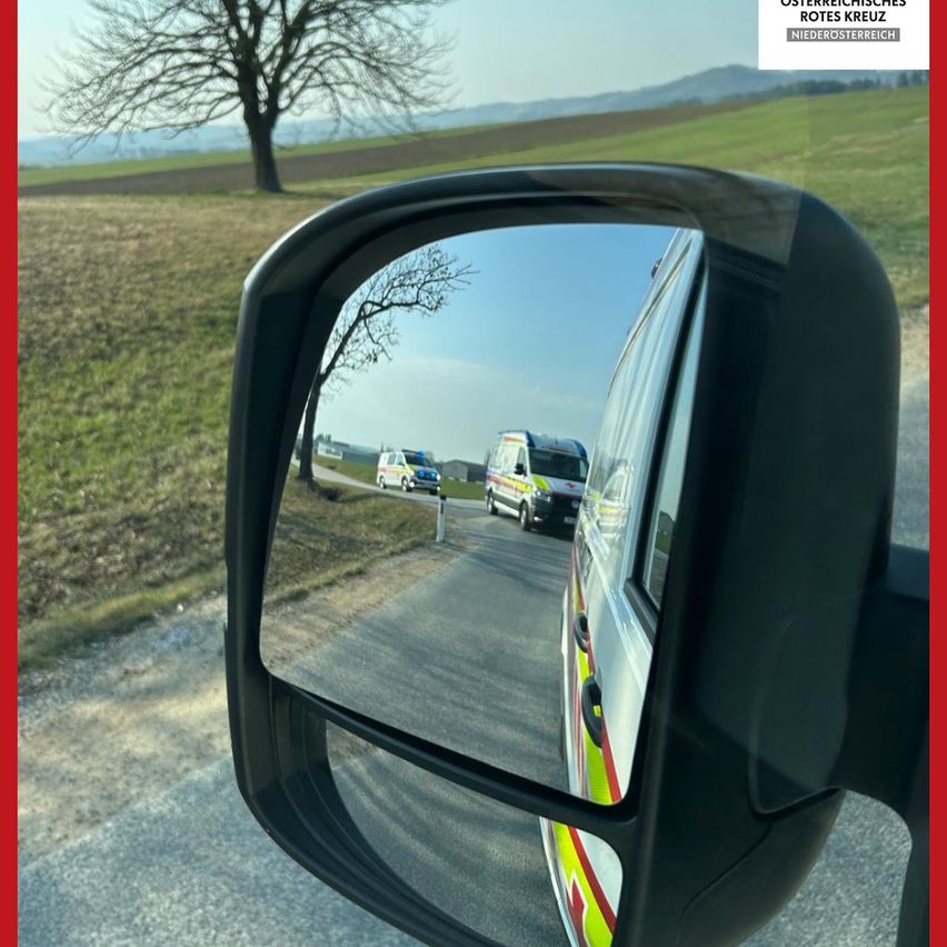 Der Rückspiegel zeigt ein Rettungsfahrzeug und zwei Polizeiautos auf einer Landstraße, mit einem Baum und grünen Feldern im Hintergrund.