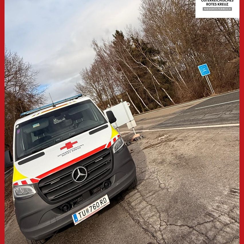 An ambulance with red cross logo parked on road with bare trees in background. The ambulance is parked with its front facing left.