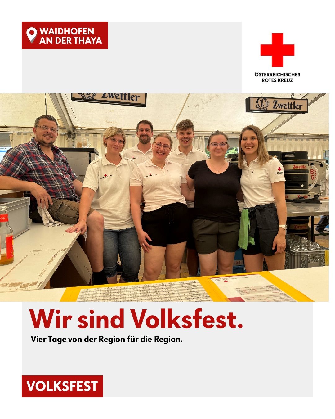 Gruppenfoto von lächelnden Freiwilligen, die weiße T-Shirts mit einem roten Kreuzlogo tragen. Sie stehen in einem Zelt mit dem Text 'Wir sind Volksfest' und 'Vier Tage von der Region für die Region.' unten.
