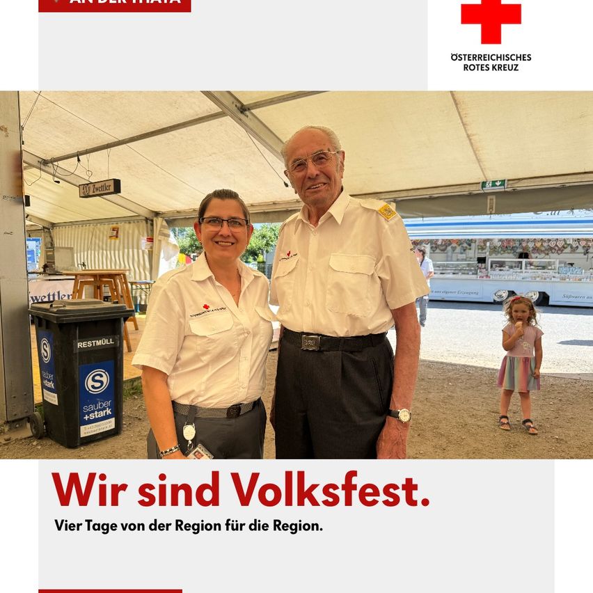 Zwei Personen, ein Mann und eine Frau, lächeln und stehen unter einem Zelt, möglicherweise bei einem Festival. Ein kleines Mädchen isst Eis. Hinter ihnen steht ein Bus, ein Mülleimer und ein Schreibtisch.