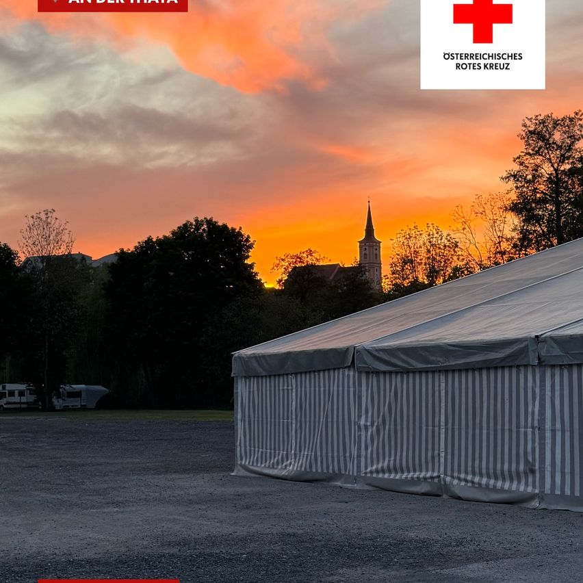 Sonnenuntergang hinter Bäumen mit einem Kirchturm, einem großen Zelt und einem Roten Kreuz Symbol.