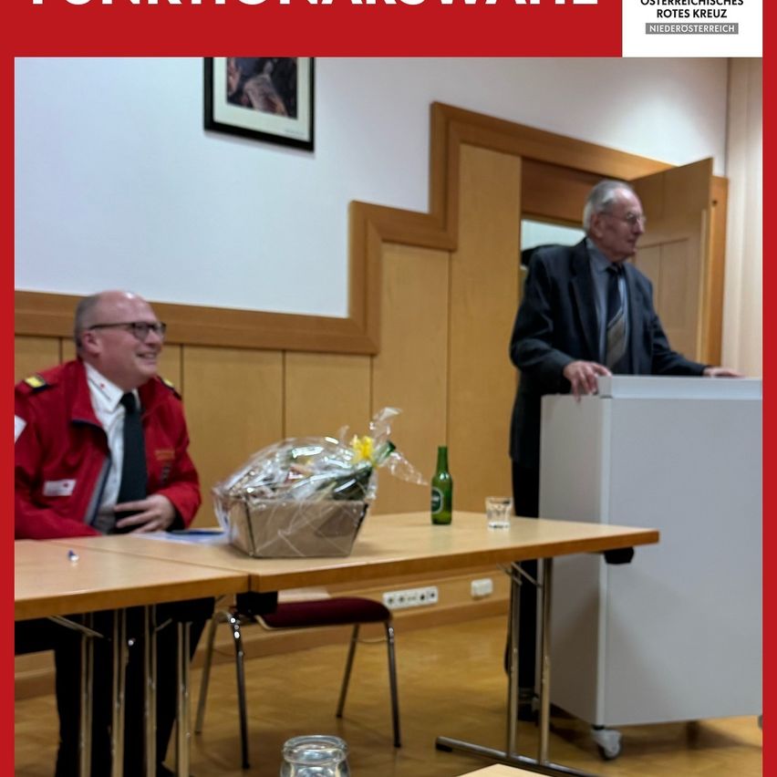 A man in a red jacket sits at a table with a bouquet and a green bottle. An older man stands at a podium, possibly giving a speech. A picture frame hangs on the wall behind him.