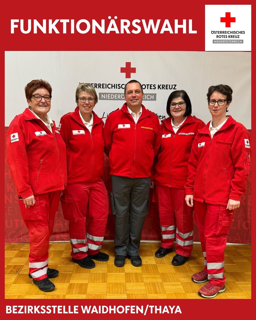 Five individuals in red jackets with white stripes stand in a row, smiling. A man in the center wears a light-colored shirt and gray pants. Behind them is a red backdrop with the text 'Rotes Kreuz Niederosterreich'.