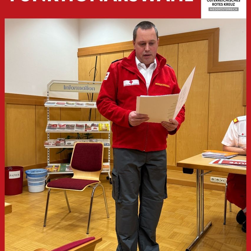 A man in a red jacket stands in a room, reading a document. Behind him, a wooden wall with a shelf holding various items. On the right, a person sits at a desk. A trash bin is on the left.