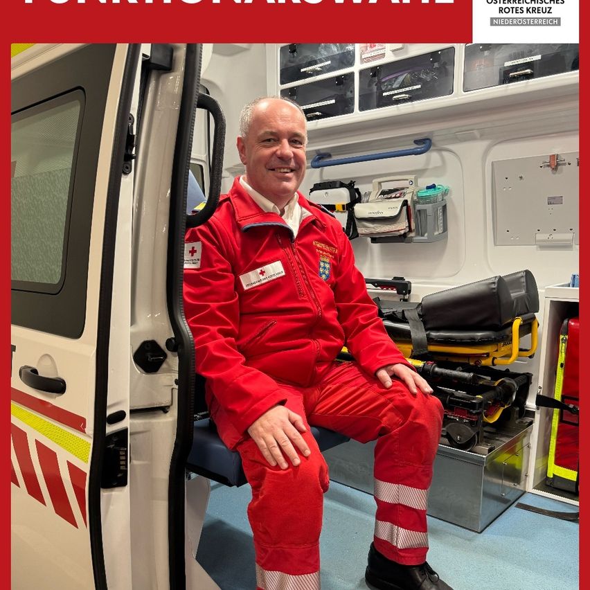 A man in a red uniform is sitting inside an ambulance, smiling. He is wearing a red jacket with a white cross emblem and red pants. Behind him is a metal tray with medical equipment.