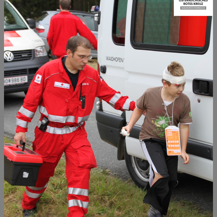 A man in red uniform helps a child with a head injury in front of an ambulance. The ambulance has red and white markings and the text 'Waidhofer' on it.