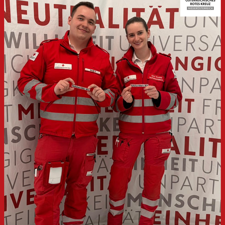Two individuals in red uniforms stand together, smiling for a photo. Both have red and white badges with text, and they are positioned in front of a red and white backdrop with blurred text.