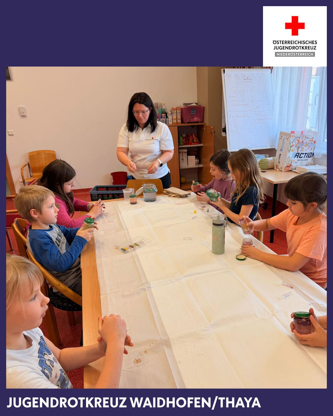 Eine Gruppe von Kindern sitzt um einen Tisch herum und führt ein Experiment durch. Eine Frau steht und erklärt ihnen etwas. Flaschen und Gläser sind auf dem Tisch.