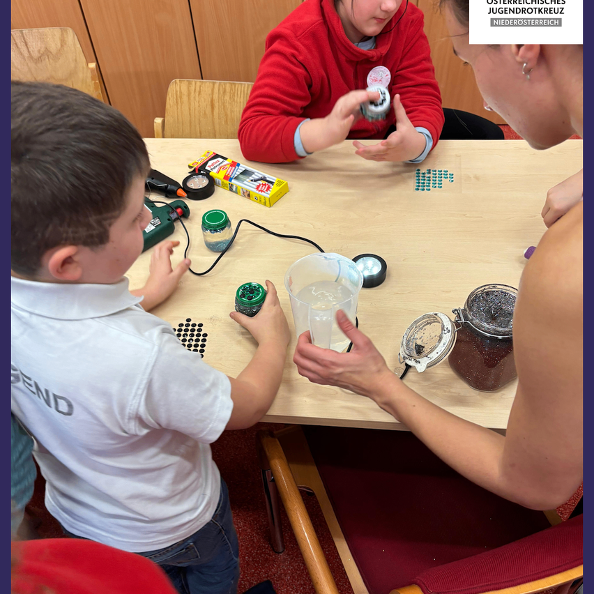 Zwei Kinder und eine Frau sind in einer Bastelaktivität engagiert. Der Junge hält ein grünes Objekt, während die Frau hilft. Der Tisch ist mit Bastelmaterialien gefüllt.