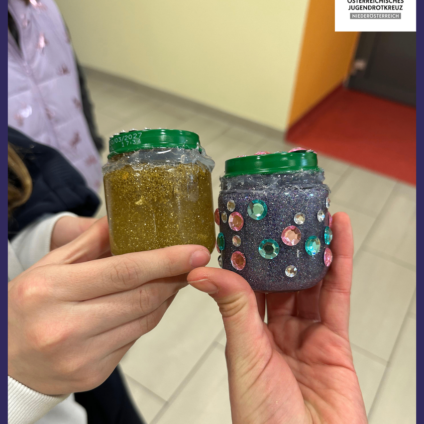 Eine Person hält zwei Gläser, eines mit glitzerndem goldenem Inhalt und eines mit glitzerndem lila Inhalt, beide mit grünen Deckeln. Der Hintergrund zeigt einen gefliesten Boden und eine Tür.