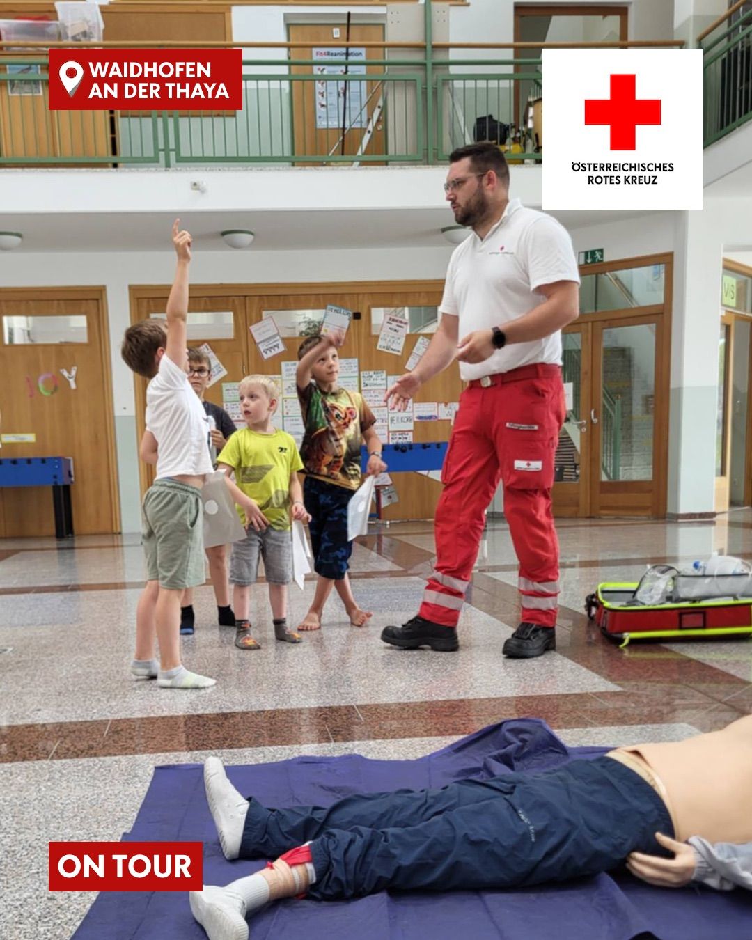Ein Mann in roten Hosen und einem weißen Hemd unterrichtet eine Gruppe von Kindern in einem Klassenzimmer. Die Kinder stehen vor ihm, und einer von ihnen hebt die Hand. In der oberen rechten Ecke befindet sich ein rotes Kreuz-Logo.