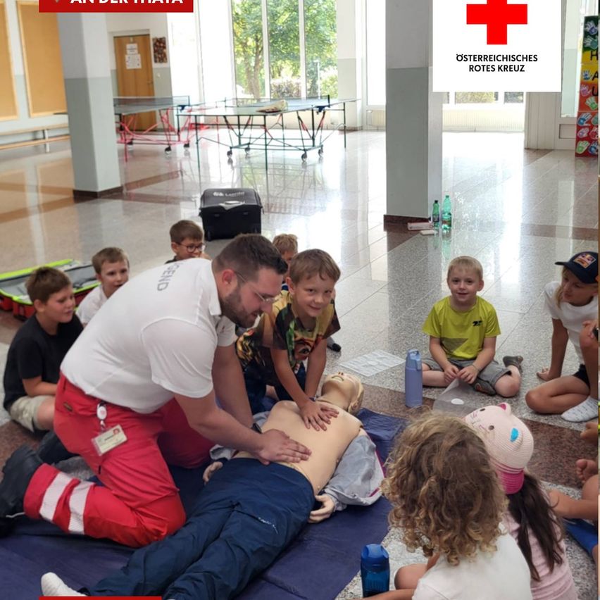 Ein Mann in einem weißen Shirt und roten Hosen demonstriert Herz-Lungen-Wiederbelebung an einem Trainingsmannequin, während Kinder aufmerksam zusehen. Der Ort ist ein Klassenzimmer mit einem roten Kreuz im Hintergrund.