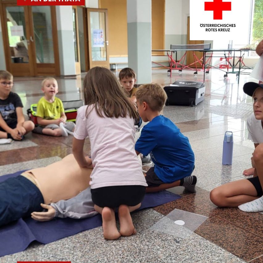 Eine Gruppe von Kindern ist um eine CPR-Puppe versammelt, wobei ein Mädchen Brustkompressionen durchführt. Andere Kinder sitzen und beobachten. Die Umgebung scheint ein Schulflur zu sein.
