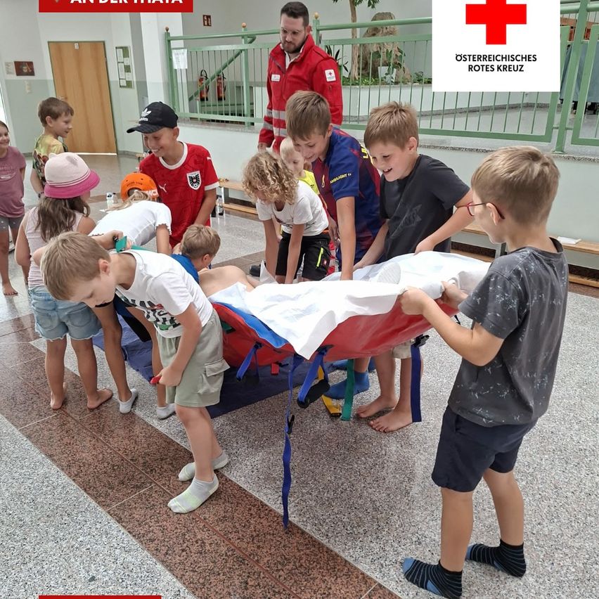 Kinder in einer Halle üben Erste Hilfe mit einem roten Kreuz im Hintergrund.