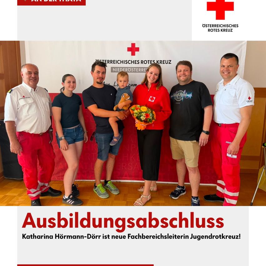 A group of people in red and white uniforms are standing together with a woman holding a bouquet of flowers and a baby. They are posing for a photo in front of a red backdrop with the text 'Ausbildungsabschluss' and the logo of the Austrian Red Cross.