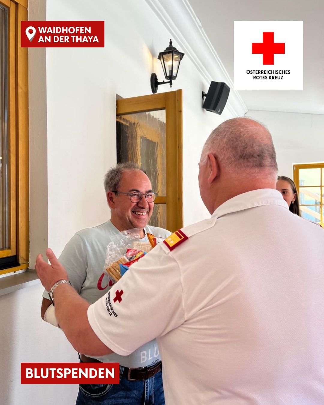 A man in a gray shirt is smiling and holding a bag of food while another man in a white shirt with a red cross emblem on his shoulder is looking at him.