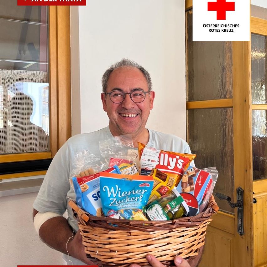 A smiling man with a bandaged arm holds a basket filled with snacks and treats, standing in front of a door with a red cross logo.