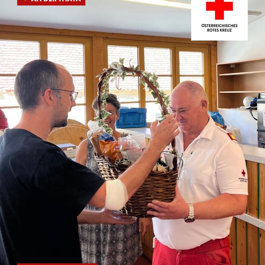 A man in a white shirt with red pants is holding a basket filled with food and other items. He is giving it to another man wearing glasses. Behind them is a woman smiling. There is a logo of the Austrian Red Cross on the top right.