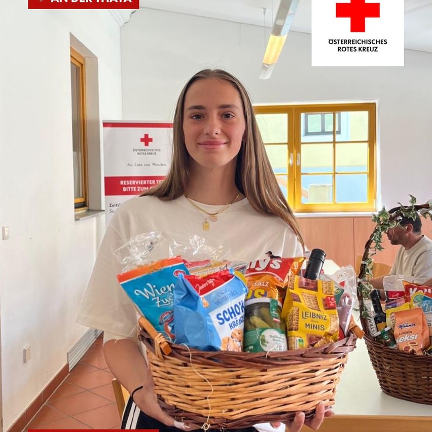 A woman holds a basket of food and snacks, smiling in front of a wall with a Red Cross sign. Another person is in the background.