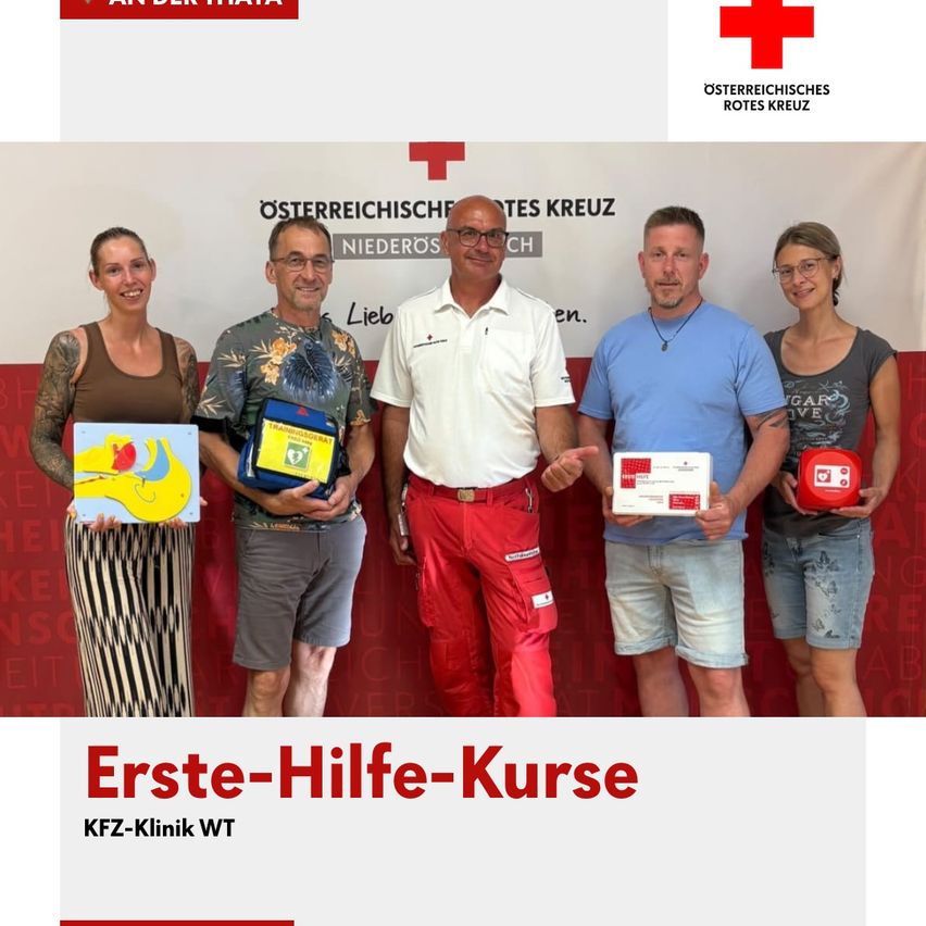 Five people pose for a photo with first aid kits and certificates. The man in the center wears a white shirt, and the others wear casual clothing. A banner with the text 'Erste-Hilfe-Kurse' and the Red Cross logo is in the background.