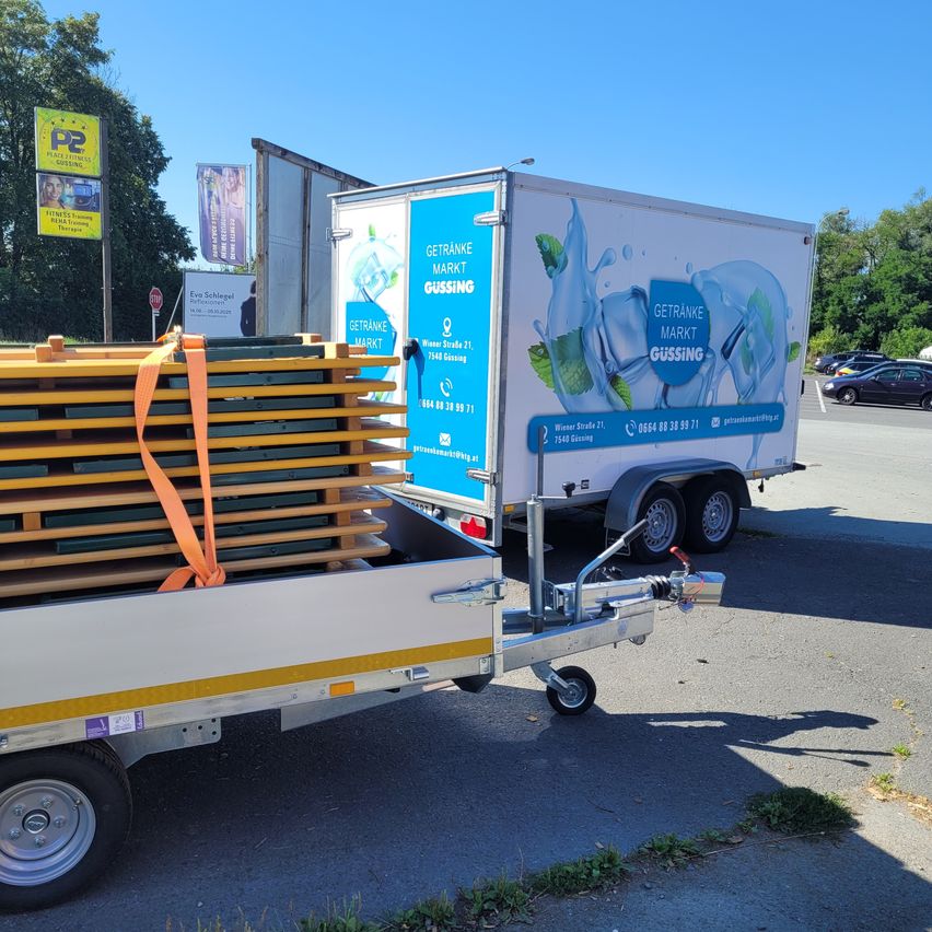 Ein weißer Anhänger ist auf der Straße geparkt, mit beladenen Holzpaletten. Er hat ein blaues Schild mit Text und Logos. In der Ferne sind Autos geparkt.
