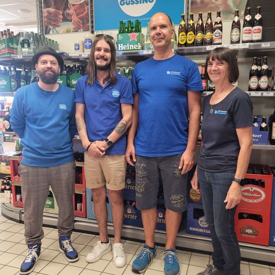 Vier Personen stehen lächelnd und posieren für ein Foto vor einem Getränkeladen. Sie tragen blaue T-Shirts und haben verschiedene Biermarken in den Regalen hinter ihnen.