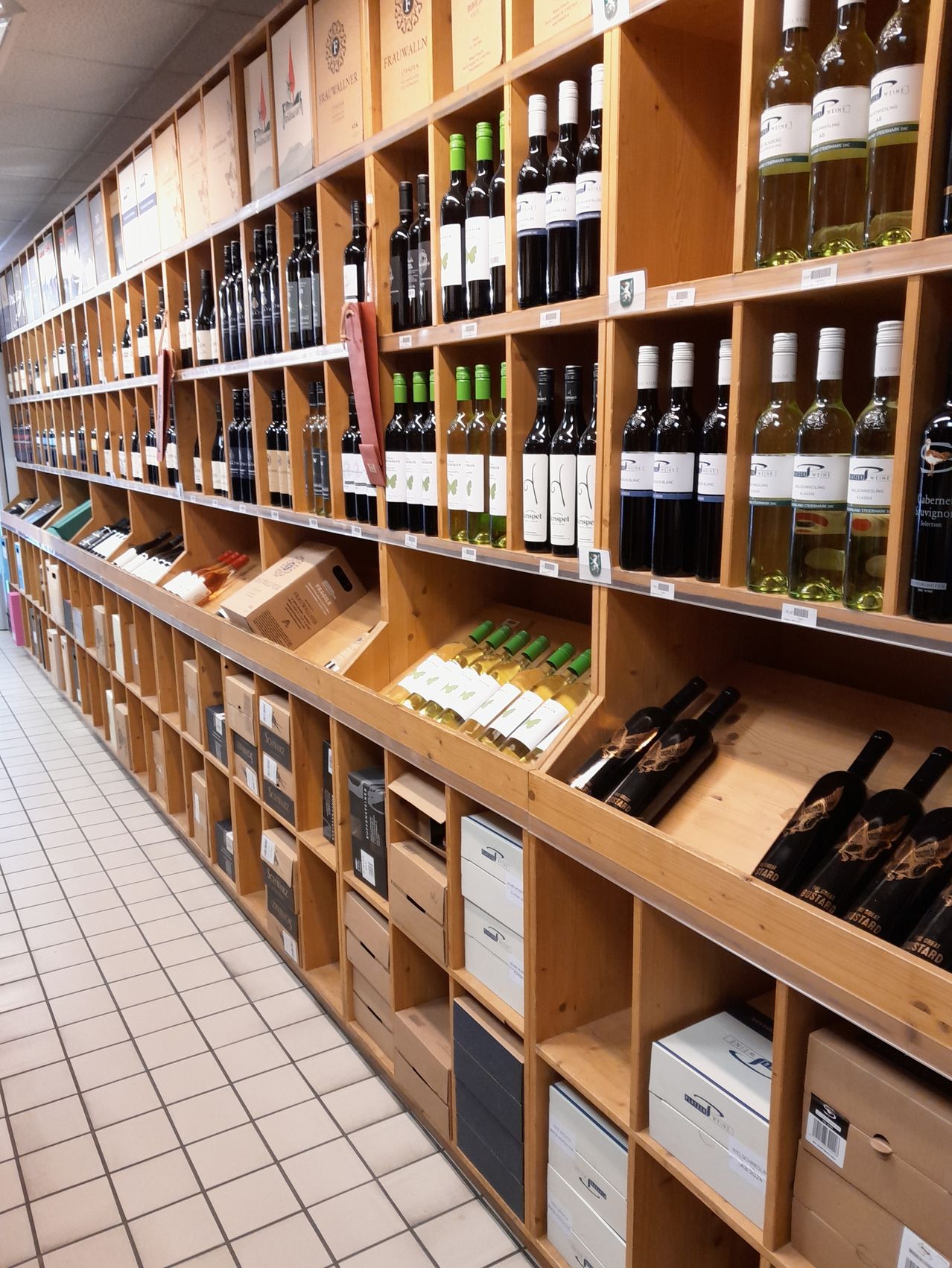 Ein Weinladen mit mehreren Holzregalen voller verschiedener Weine, einige mit Etiketten, andere ohne. Der Boden ist weiß gefliest.