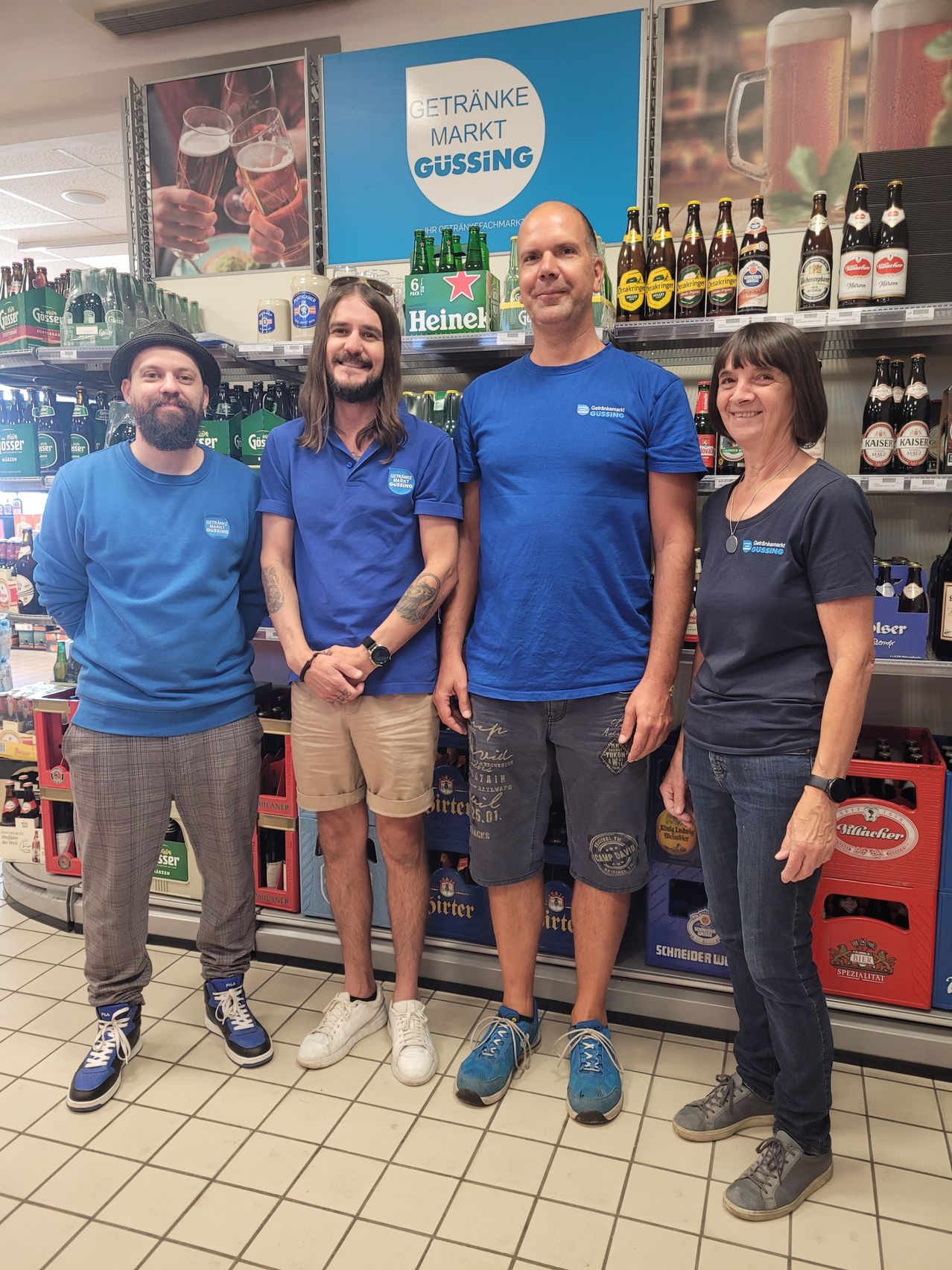 Vier Personen stehen in einem Bierladen mit blauen T-Shirts und lächelnden Gesichtern. Hinter ihnen sind Regale mit Bierflaschen. Einer von ihnen hat ein Tattoo auf dem Arm.