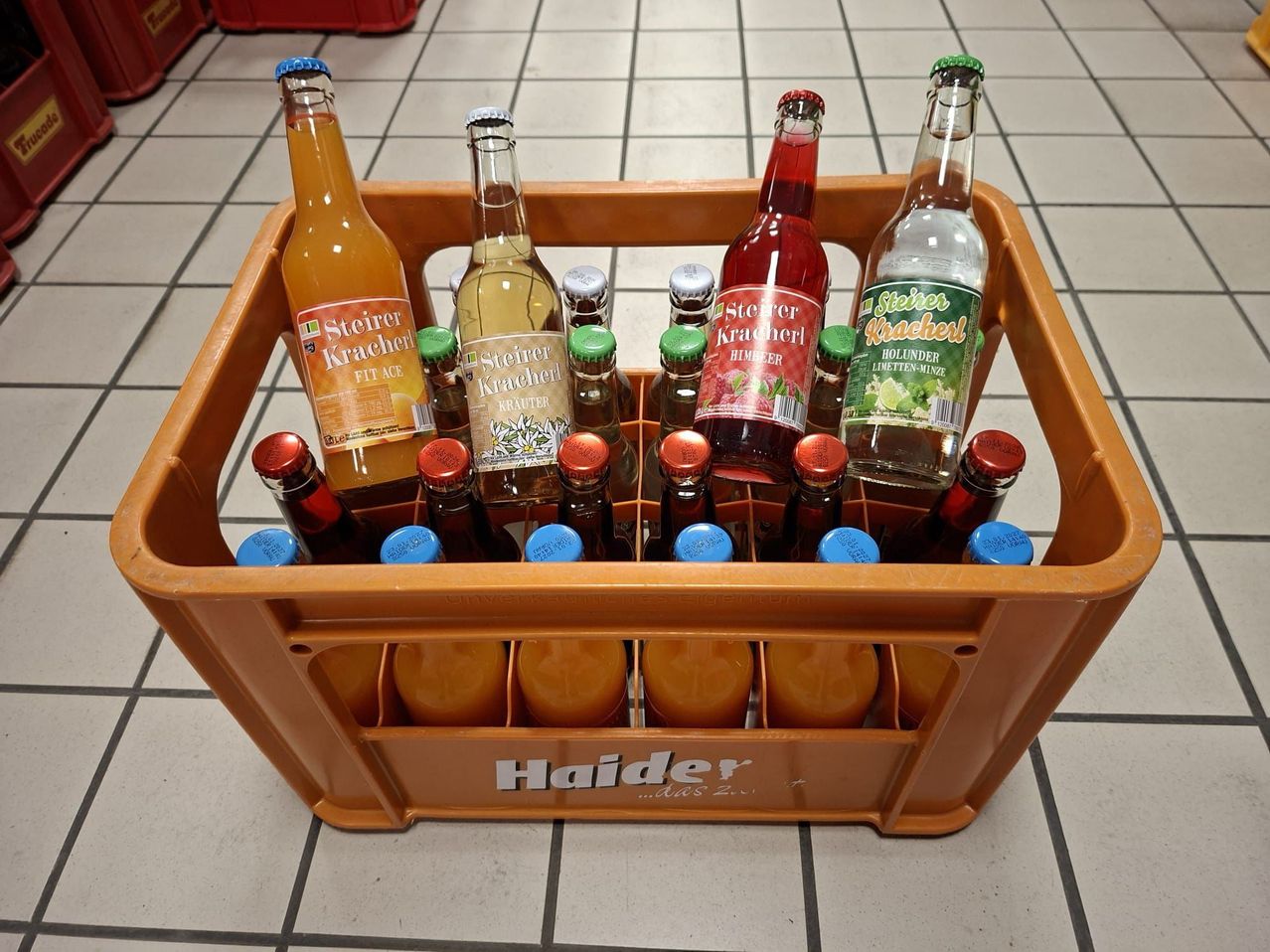 Ein orangefarbener Kasten mit der Aufschrift Haider enthält mehrere Flaschen Steiraer Kracher und Hollunder Limetten-Muntz. Die Flaschen sind in zwei Reihen angeordnet.