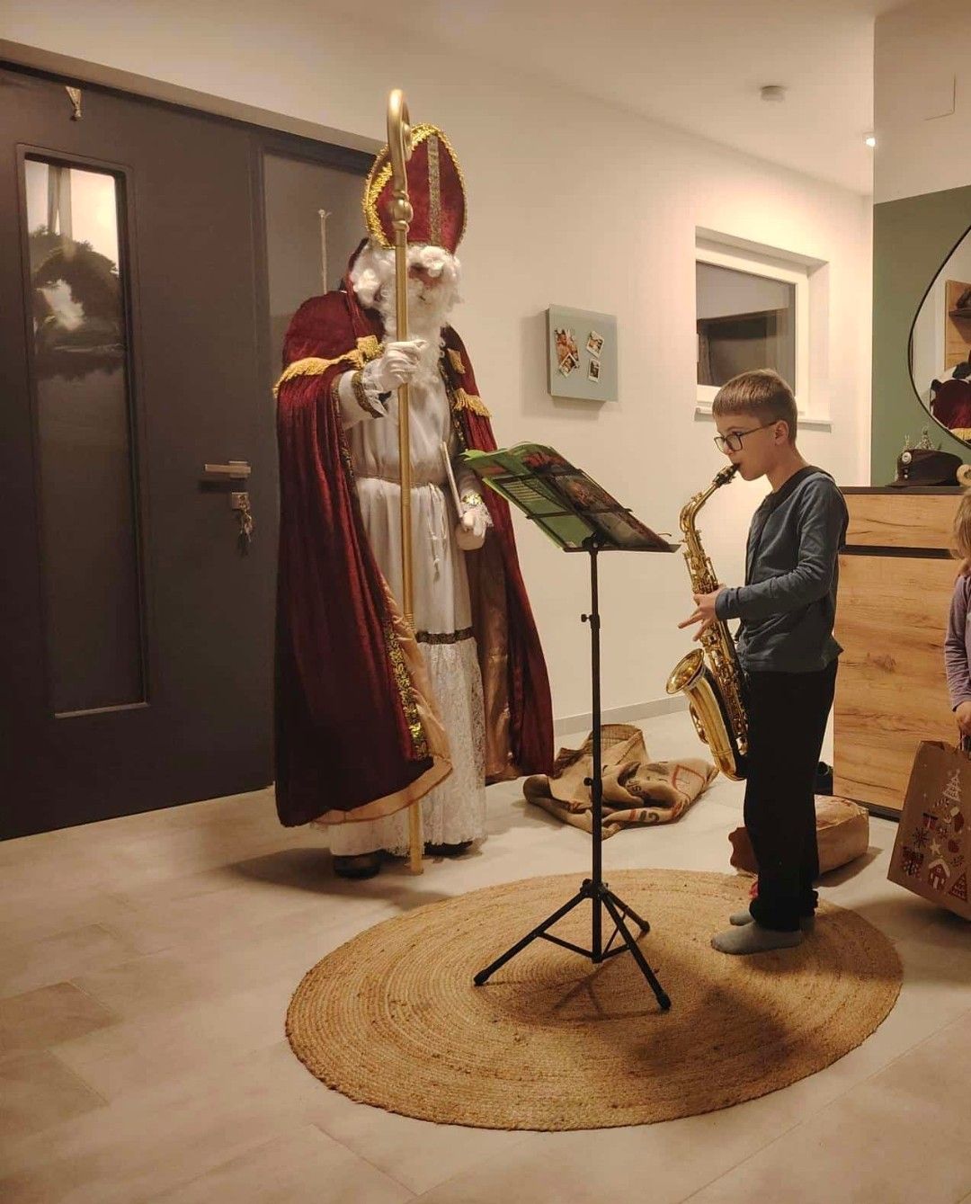 Ein Junge spielt die Saxophon, während der als Weihnachtsmann verkleidete Mann hinter ihm steht. Ein Mädchen mit einem Geschenkbeutel steht rechts, und ein Kranz hängt an der Tür.