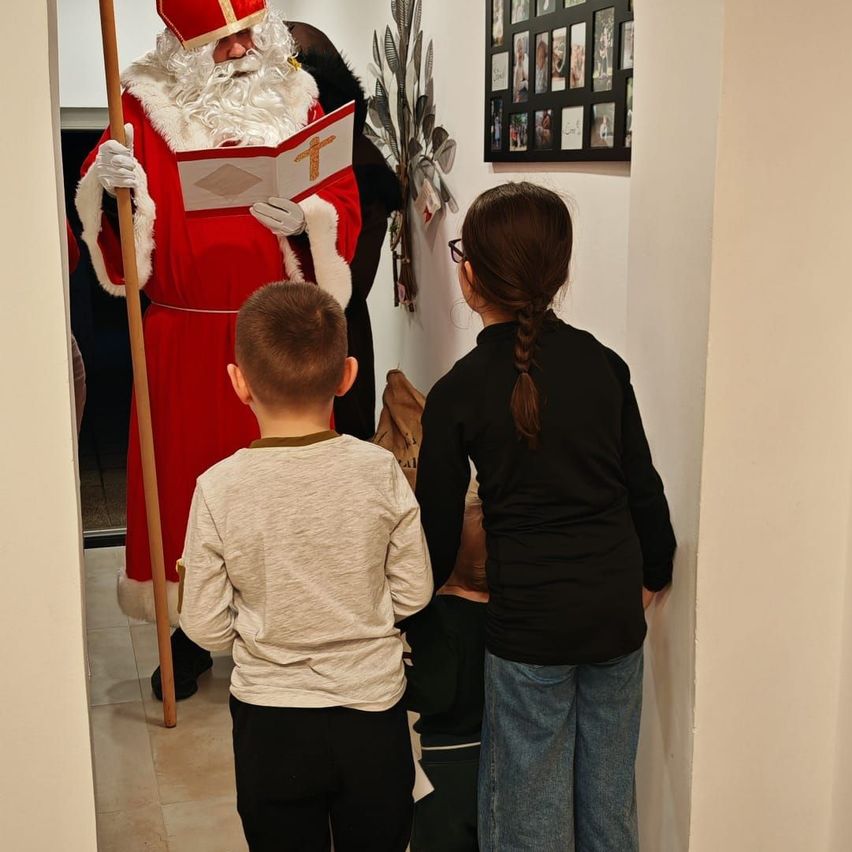 Ein Mann in Santa Claus-Kostüm hält ein Buch mit einem Kreuz, während ein Junge und ein Mädchen aus dem Flur zusehen.