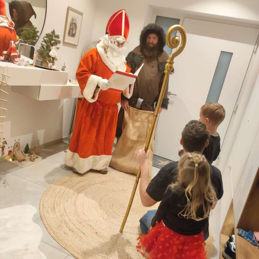 Eine als Weihnachtsmann verkleidete Person liest Kindern ein Buch vor, während ein als Elf verkleideter Mann hinter ihr einen Stab hält, in einem Raum.