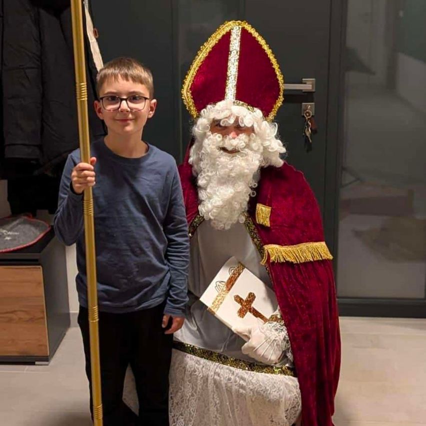 Ein Junge mit Brille hält einen goldenen Stab in der Hand und steht neben einer Person, die als St. Nikolaus in einem roten Gewand und Hut gekleidet ist.