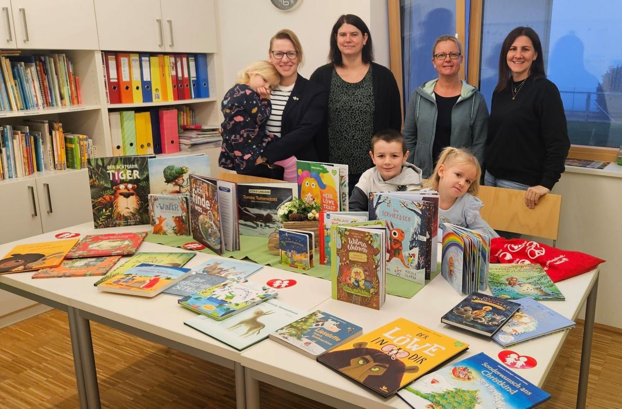 Eine Gruppe von Erwachsenen und Kindern steht hinter einem Tisch voller verschiedener Kinderbücher, mit einem Regal und einer Uhr im Hintergrund.