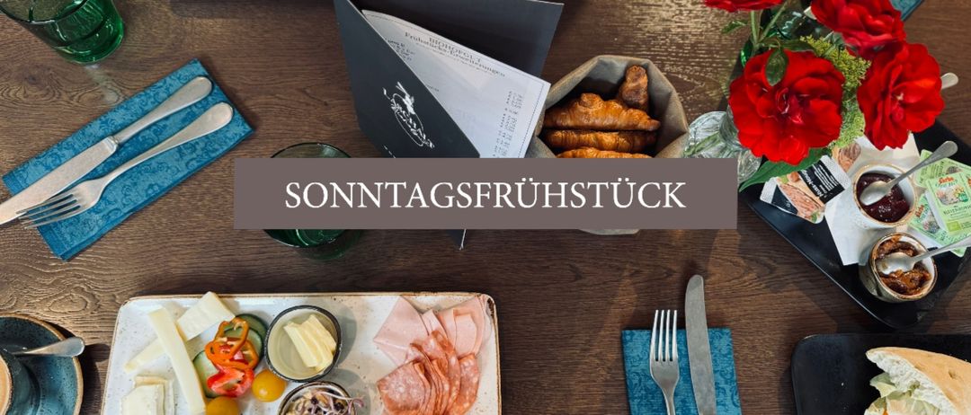 Ein Holztisch zeigt verschiedene Essensplatten mit Fleisch, Käse und Gemüse. Es gibt auch Blumen, Besteck und ein Menübuch. Der Text 'SONNTAGSFRUHSTUCK' steht auf einem grauen Hintergrund.