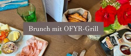Ein Brunchtisch mit einem hölzernen Menübuch, einer Schüssel mit Croissants und einem Teller mit Fleisch. Der Tisch ist mit Blumen und einem grünen Glas dekoriert.