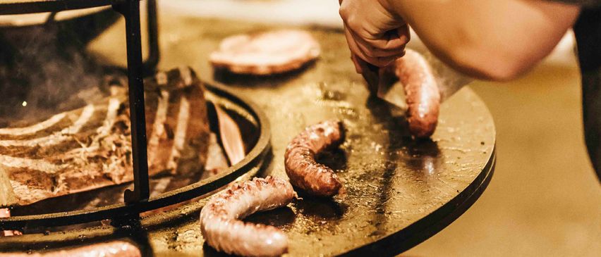 Eine Person grillt Würste und Fleisch auf einem Grill mit dem Text 'GRILLKURS' darüber.
