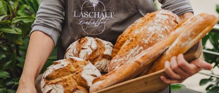 Eine lächelnde Frau in einer grauen Schürze mit LASCHALT Biohofgut darauf hält eine Tablett mit frisch gebackenem Brot. Der Hintergrund ist ein Garten mit grünen Bäumen.