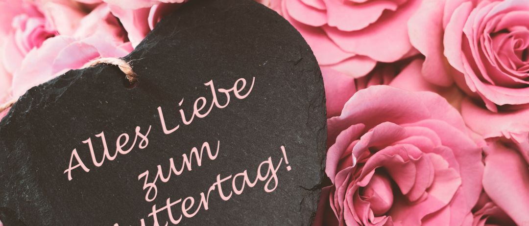 Ein Herz mit einer Nachricht, die in rosa Tinte auf einem dunklen Hintergrund geschrieben ist, mit rosa Blumen im Hintergrund. Die Nachricht lautet 'Alles Liebe zum Muttertag!' auf Deutsch.