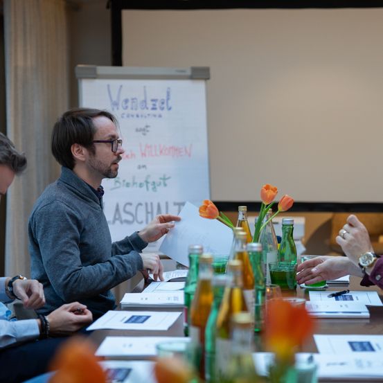 A group of people are gathered around a table with various items. One man is speaking while another listens. A whiteboard with the word 'Wendzel' is visible in the background.