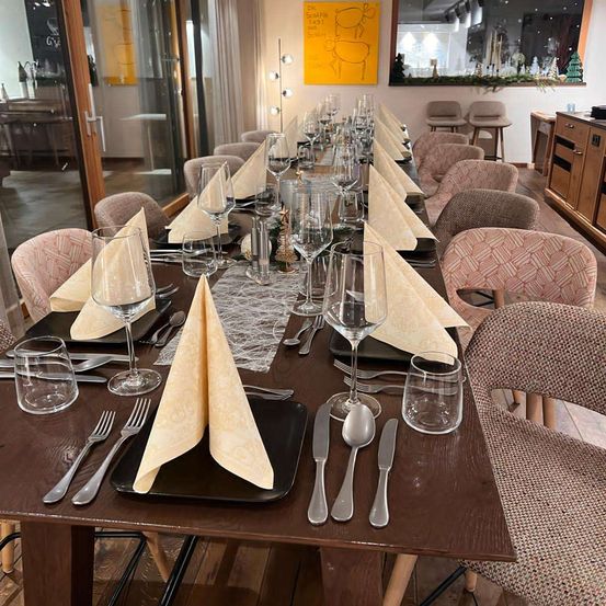 A long dining table is set for a meal. The table is adorned with wine glasses, napkins, and silverware on black plates. The room is warmly lit, and there is a Christmas decoration on the wall.