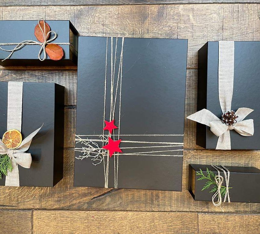 Fünf schwarze Geschenkboxen liegen auf einer Holzoberfläche. Jede ist in weiße Bänder eingewickelt. Die mittlere Box hat rote Sterne.