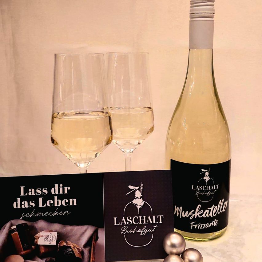 Zwei mit Weißwein gefüllte Weingläser stehen neben einer Flasche Weißwein mit dem Etikett 'Muskatelle Frizzante.' Eine schwarze Karte mit 'LASS DICH DAS LEBEN SCHMECKEN' und 'LASCHALT BISCHHOFGUT' ist ebenfalls vorhanden.
