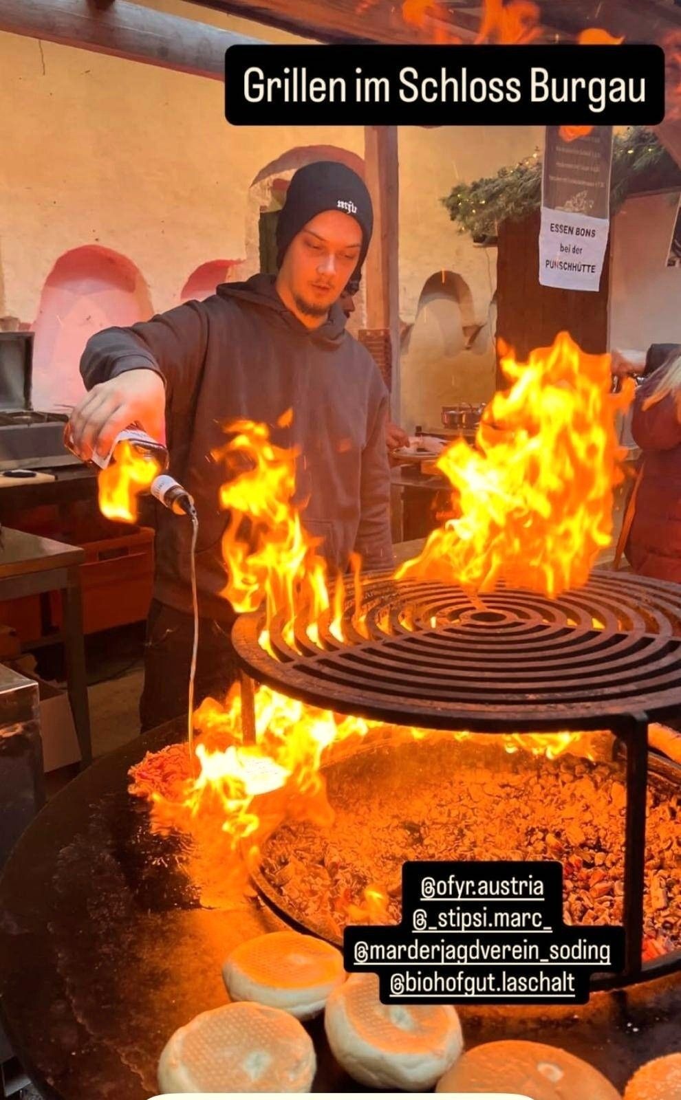 Ein Mann gießt eine brennbare Flüssigkeit auf einen brennenden Teller, mit Zuschauern in der Nähe. Ein Schild steht 'Essen Bons bei der Punschhütte'.