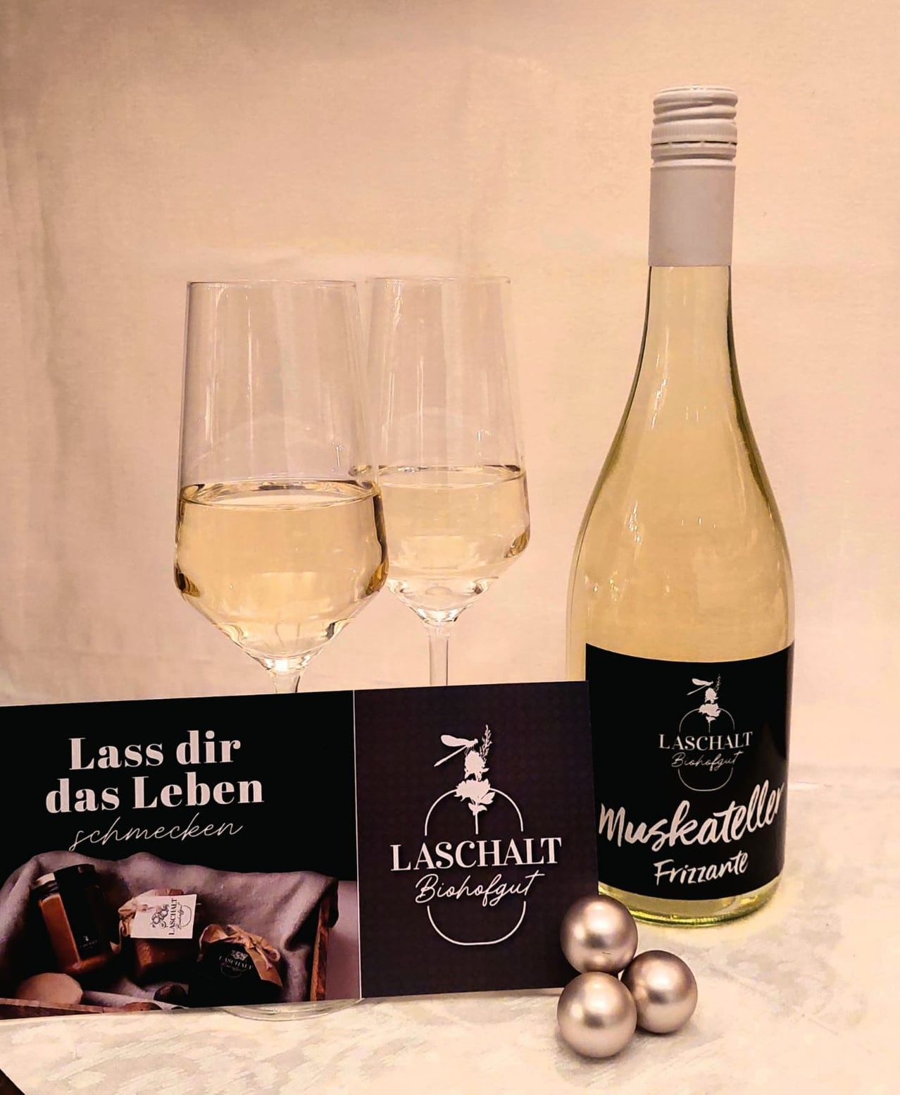 Zwei mit Weißwein gefüllte Weingläser, eine Flasche Weißwein und eine Geschenkbox mit verschiedenen Artikeln. Die Weinflasche ist mit 'Muskateller Frizzante' beschriftet. Die Weingläser und die Geschenkbox sind auf einer Marmoroberfläche platziert.